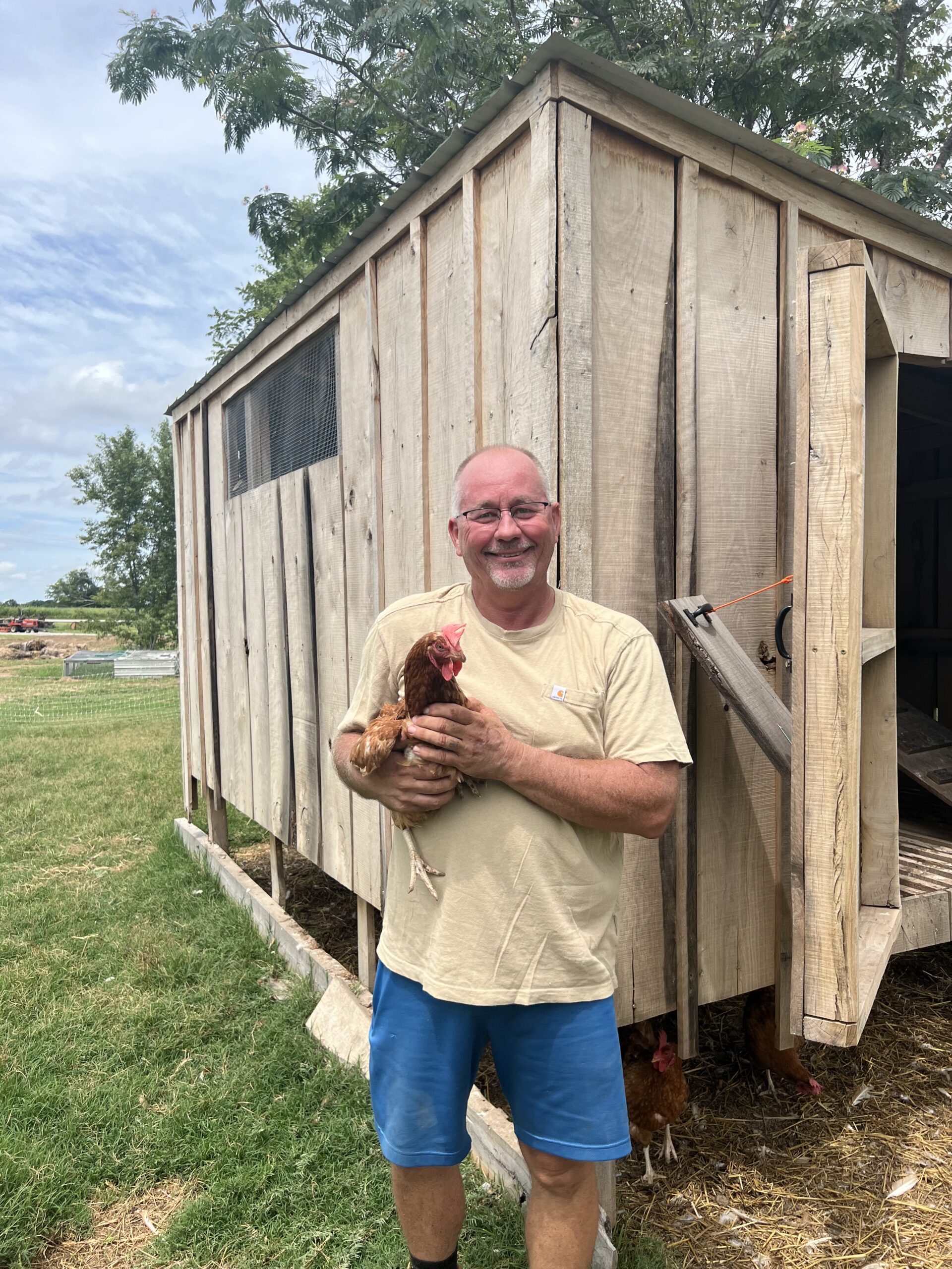 A farmer holding a chicken stands in front of a wooden chicken coop surrounded by green grass and trees, showcasing sustainable poultry farming practices.