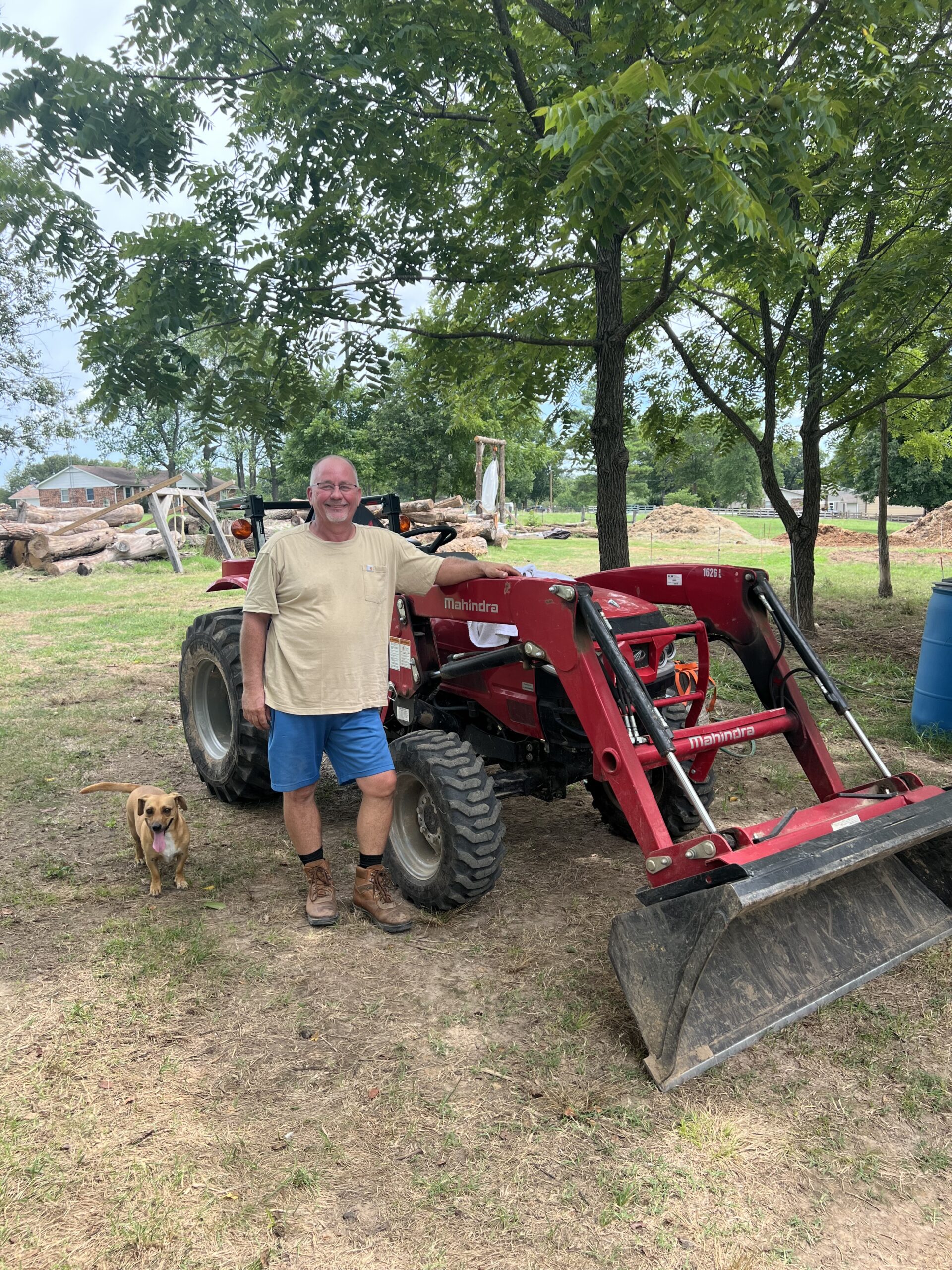 Man standing next to a red Mahindra tractor in a rural setting, with a brown dog nearby, surrounded by trees and a background of logs and earth.