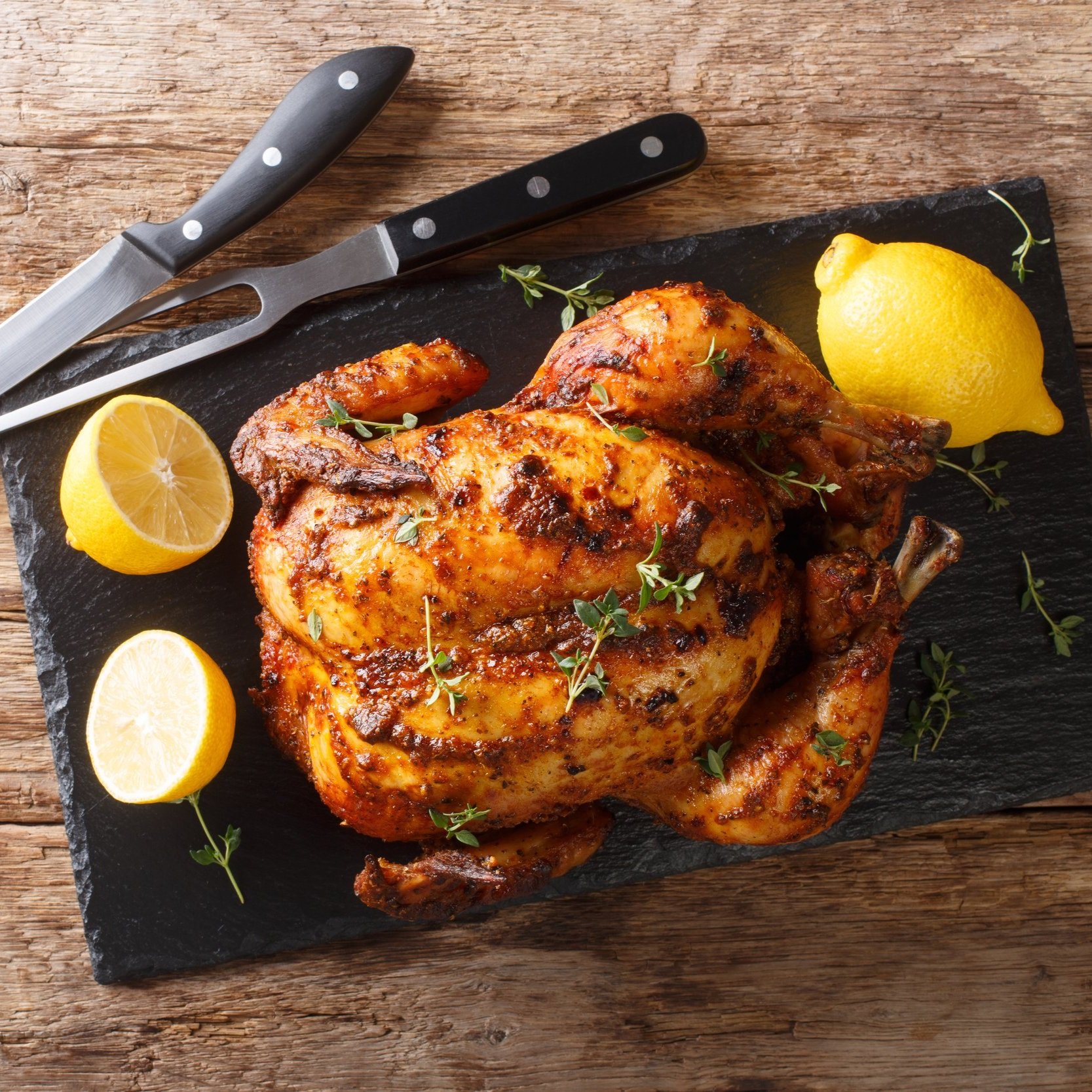 Roast chicken garnished with fresh thyme and served with lemon wedges on a black slate platter, accompanied by sharp kitchen knives on a rustic wooden table.