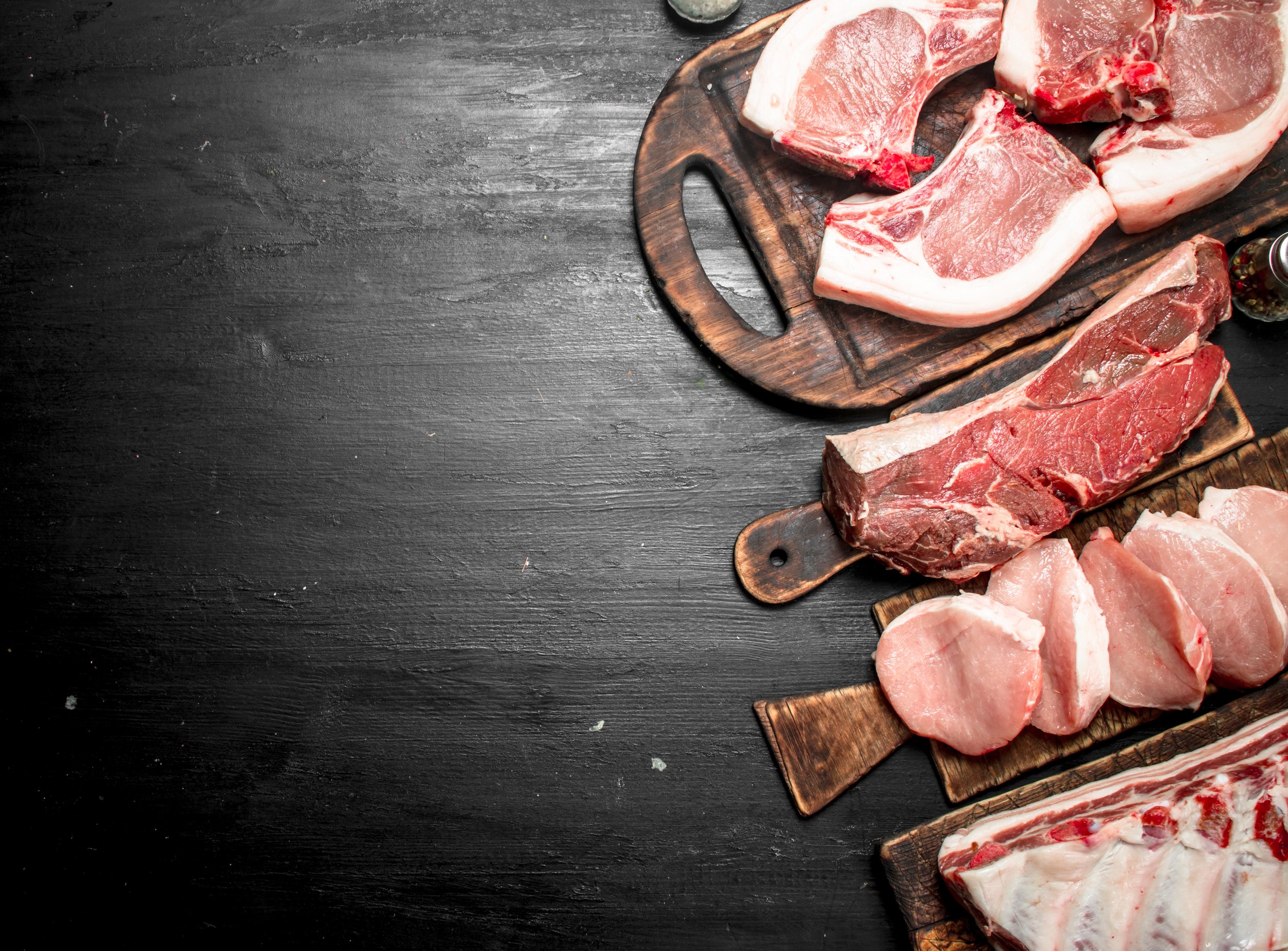 Assorted cuts of raw meat displayed on rustic wooden cutting boards, including pork chops, beef ribeye, and sliced chicken breast, set against a dark textured background.