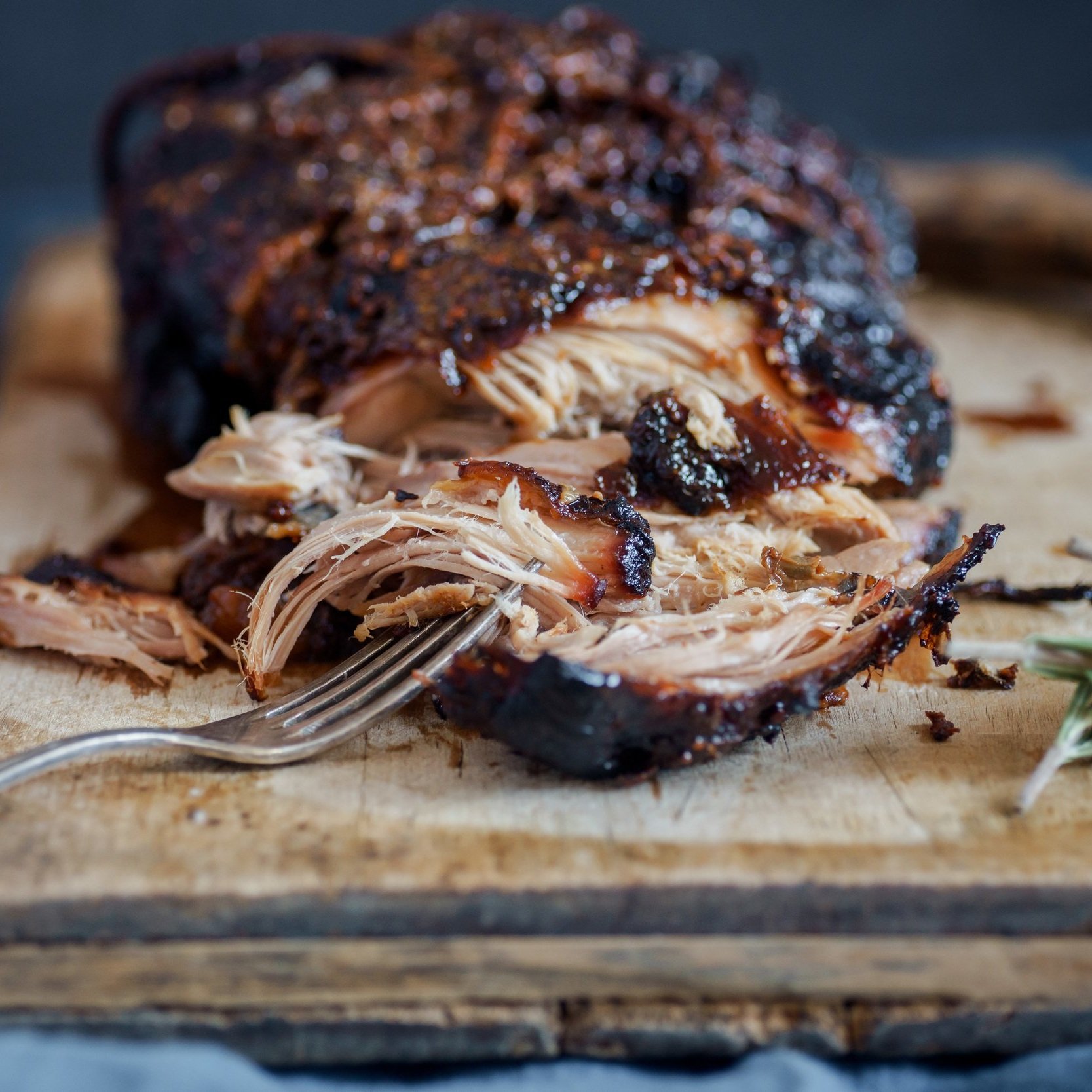 Smoked pulled pork on a wooden cutting board, showcasing tender, shredded meat with a fork resting beside it, highlighting its rich, caramelized exterior and juicy texture.